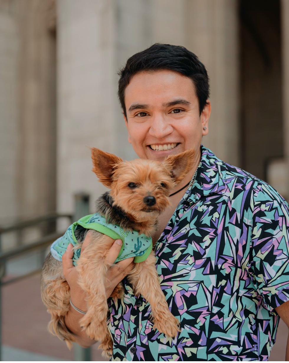 Reggie Casanova-Perez smiling while holding a small dog, wearing a colorful patterned shirt